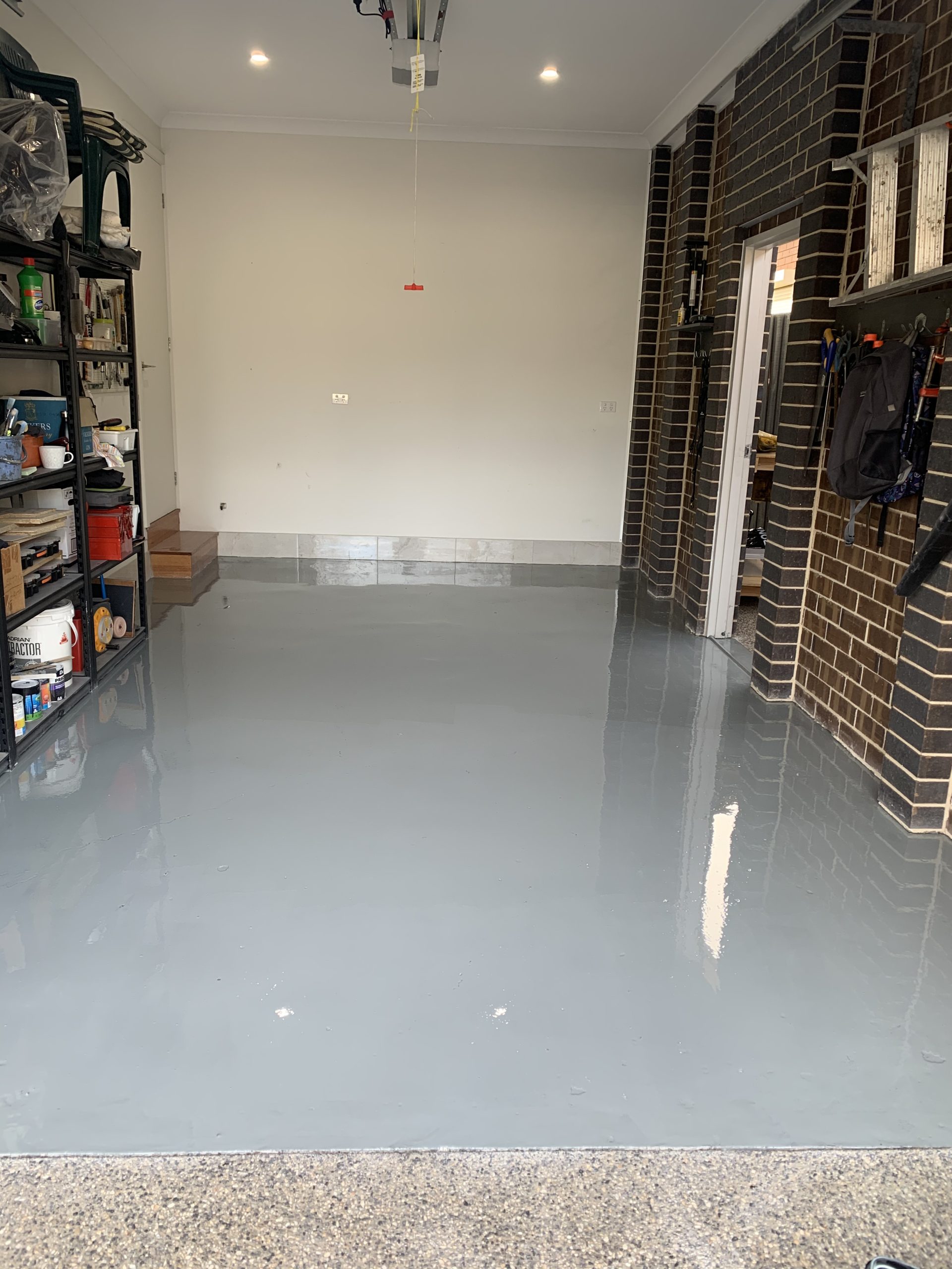 High-gloss grey epoxy floor coating in brick home garage with shelving and exposed aggregate driveway