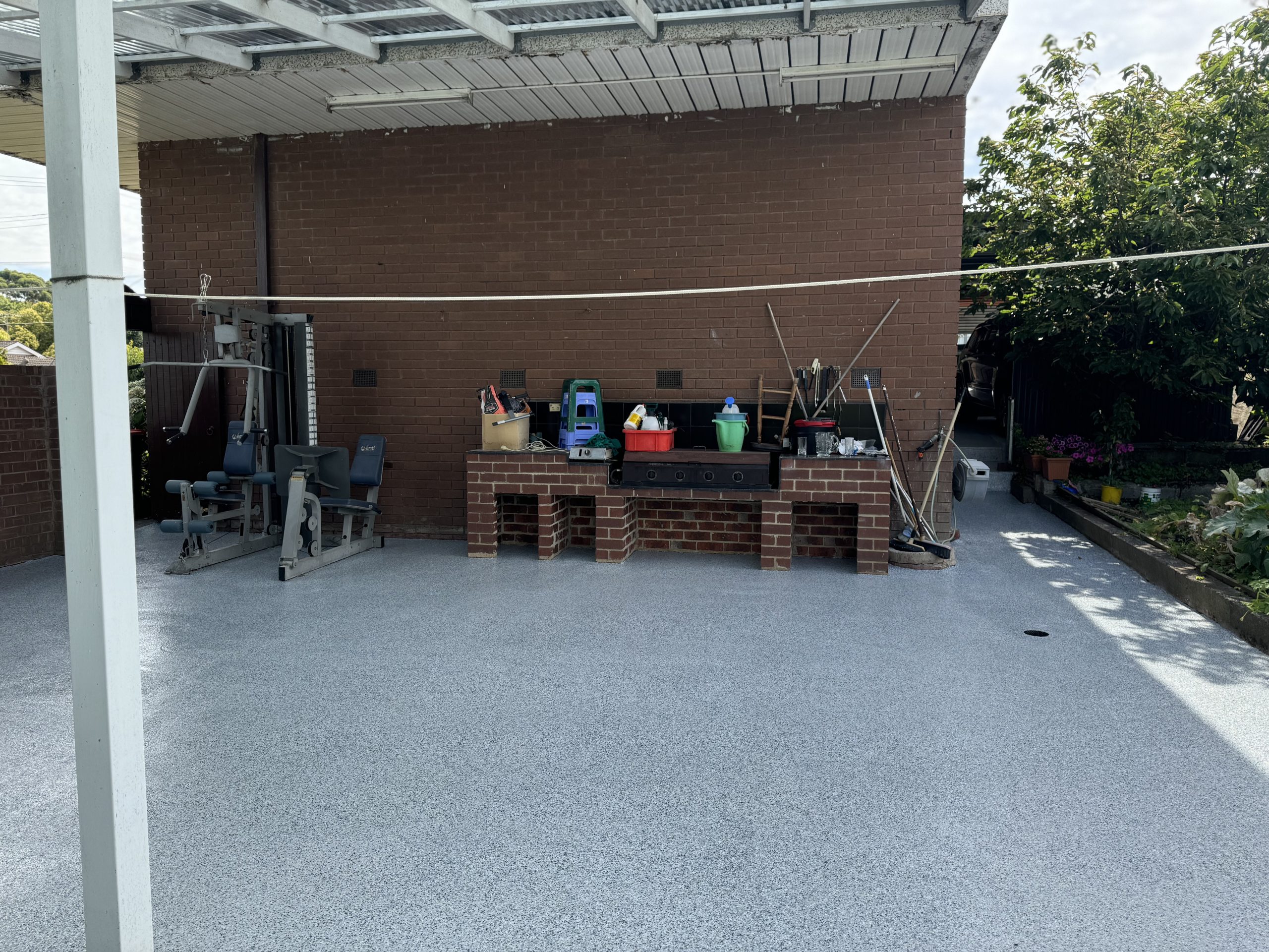 Covered outdoor area with light grey epoxy flake coating, brick BBQ, gym equipment, and garden surrounds