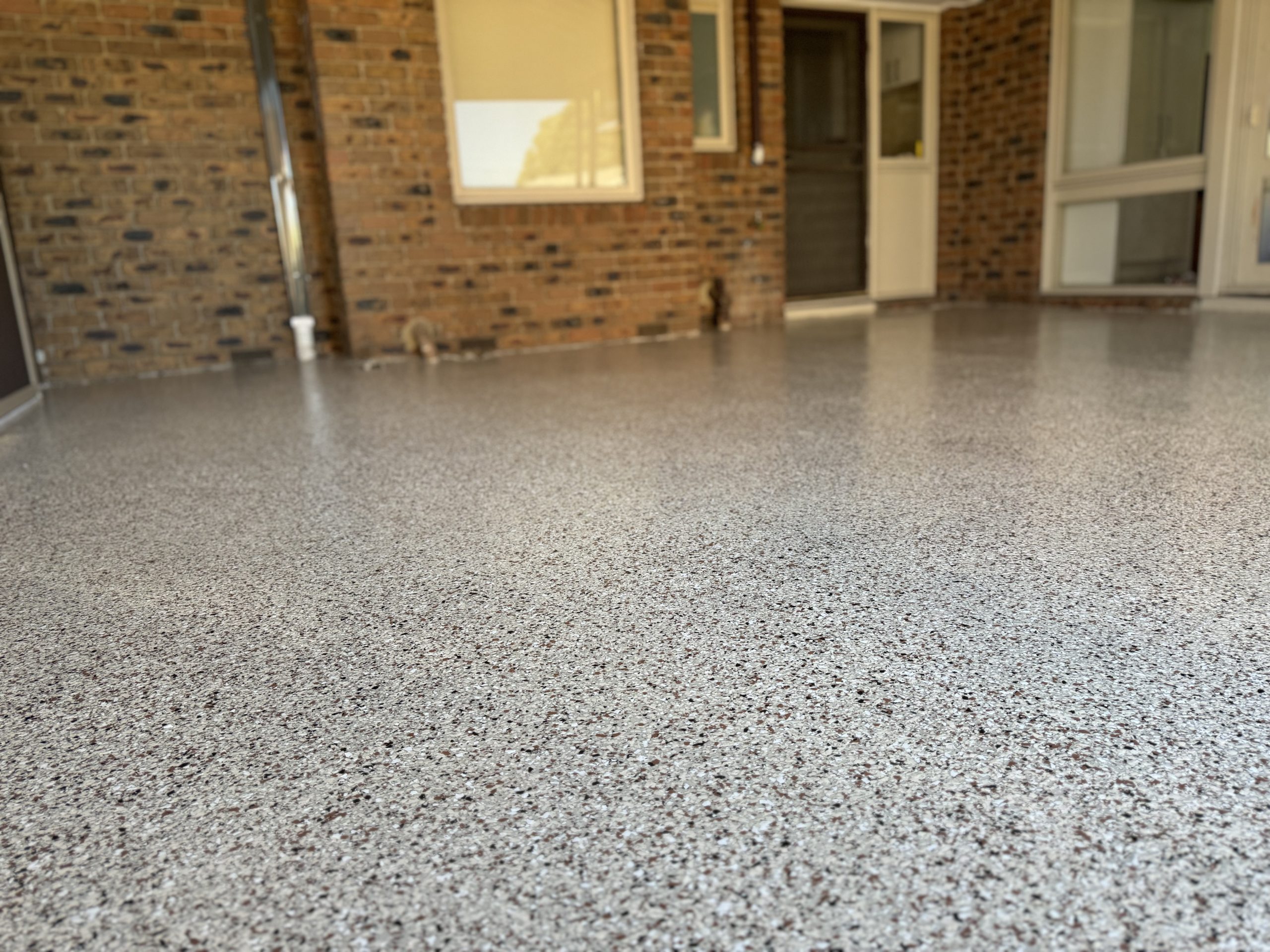 Close-up of grey and brown epoxy flake garage floor coating with brick home exterior and screen door in background